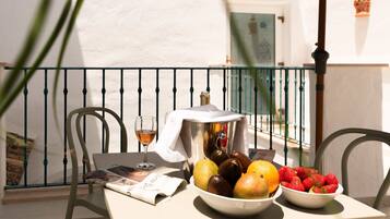A table with a newspaper, a glass of wine, a bowl of fruit, and a metal container on a balcony with a railing.