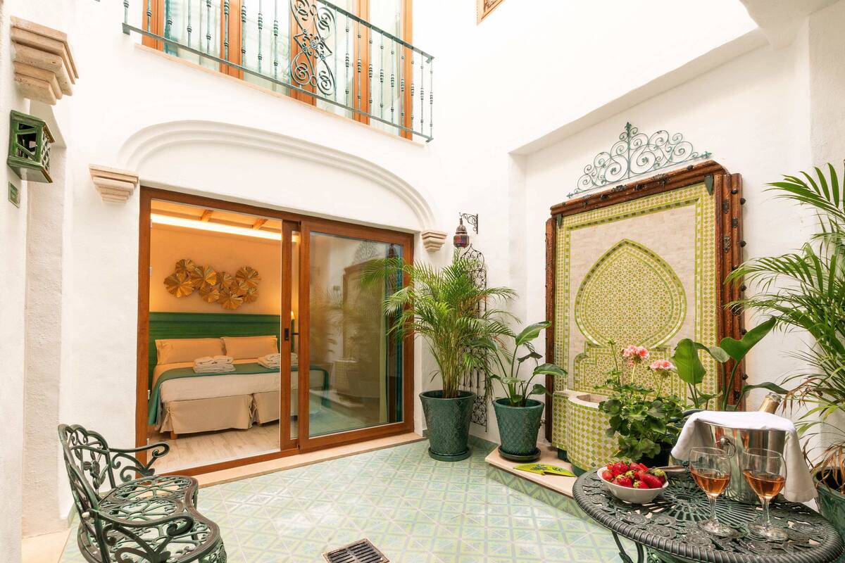 A courtyard with a bed, potted plants, a table with glasses, and a decorative door.