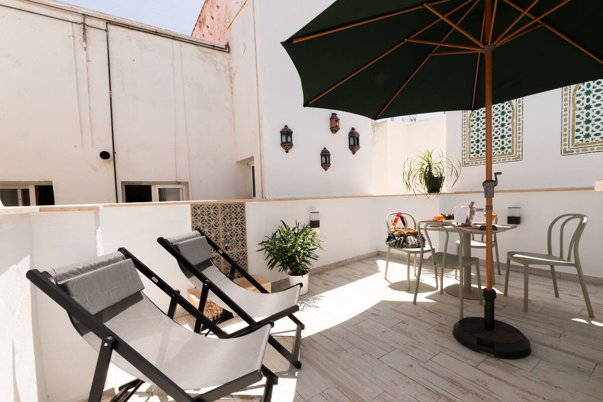A rooftop patio with lounge chairs, a table with chairs, a potted plant, and decorative wall lanterns.