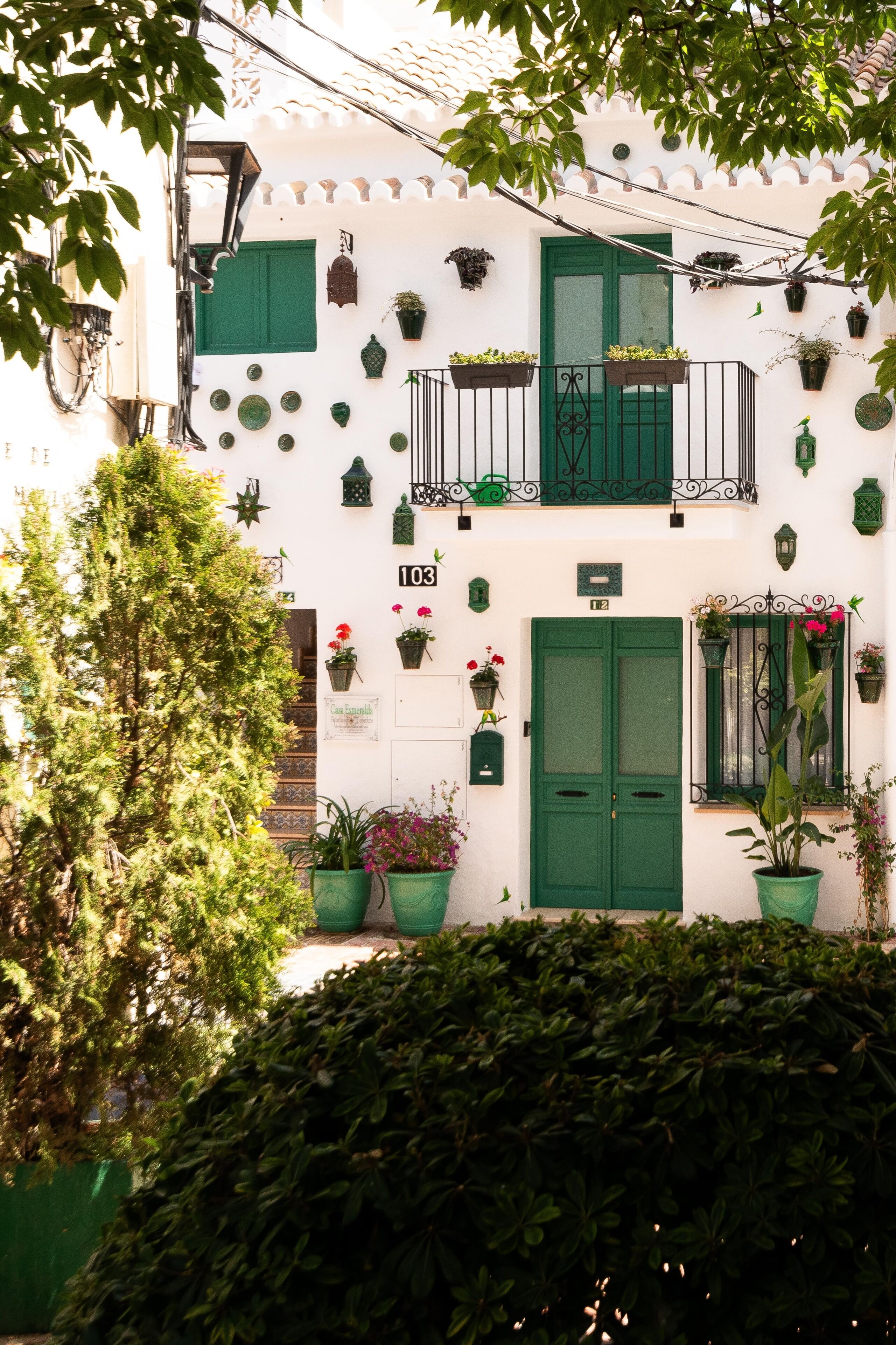 A white building with green shutters and balconies, adorned with potted plants and flowers.