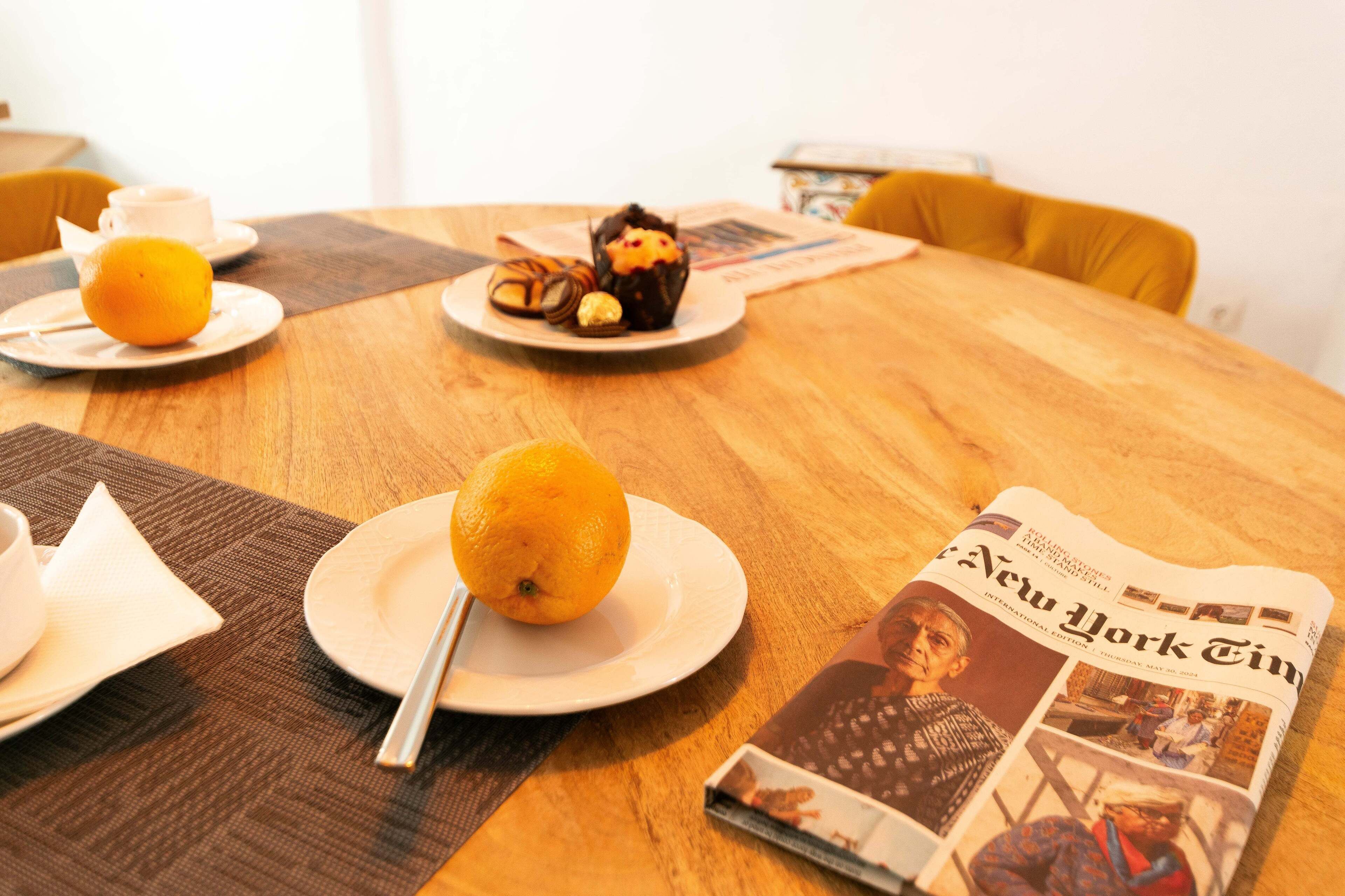 A wooden table with a newspaper, a plate with an orange, and a plate with pastries.