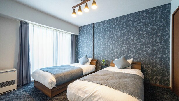 Ein Hotelzimmer mit zwei Betten, einem großen Fenster mit Vorhängen, einer blumengemusterten Wand und einer Deckenleuchte.