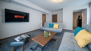 Ein modernes Wohnzimmer mit einem Flachbildfernseher, der 'NETFLIX' zeigt, einem grauen Sofa mit blauen und gelben Kissen, einem Holztisch mit einer Zeitschrift und einer grauen Couch mit einer gelben Decke.