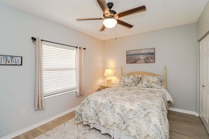 Um quarto com cama, um ventilador de teto, uma mesa de cabeceira com abajur, uma janela com persianas e um quadro na parede.