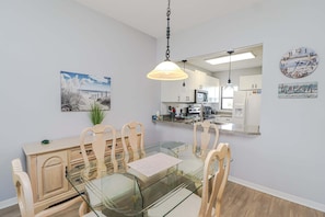 A dining area with a glass table, wooden chairs, a hanging light fixture, a kitchen in the background, and a beach-themed wall clock.