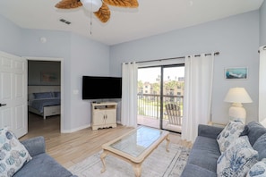 A modern living room with a flat-screen TV, a glass-top coffee table, a sofa, and a balcony with a view.