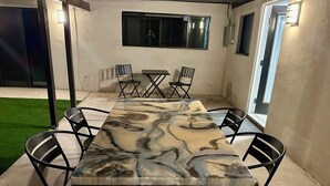Une terrasse extérieure avec une table rectangulaire et six chaises, une petite fenêtre et une porte donnant sur un autre espace.