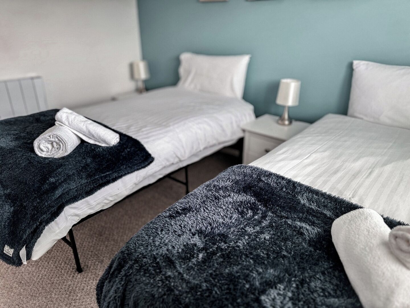 Et hotelværelse med to enkeltsenge, hver med et hvidt dynebetræk og et mørkegråt tæppe. Der er sengeborde med lamper, og væggene er malet i en lys blå farve.