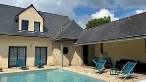Una piscina con sillones de descanso, un muro de piedra y un edificio de dos plantas con techo oscuro.