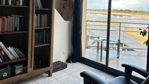 Una habitación con una amplia estantería, una silla de oficina negra y vista a una playa a través de un gran ventanal.