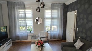 Ein Wohnzimmer mit einer Couch, einem Couchtisch mit Blumen und einem Fernseher auf einem Schrank sowie einem Sessel.
