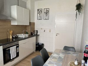Eine moderne Küche mit weißen Schränken, einer schwarzen Arbeitsplatte und einem Edelstahlbackofen. An der Wand hängen zwei Bilder, eine Hängepflanze und ein Esstisch mit Stühlen.