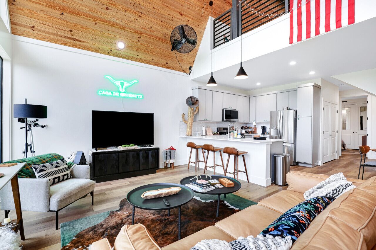 A modern living room with a flat-screen TV, a sofa, a coffee table, and a kitchen area with white cabinets and a bar.