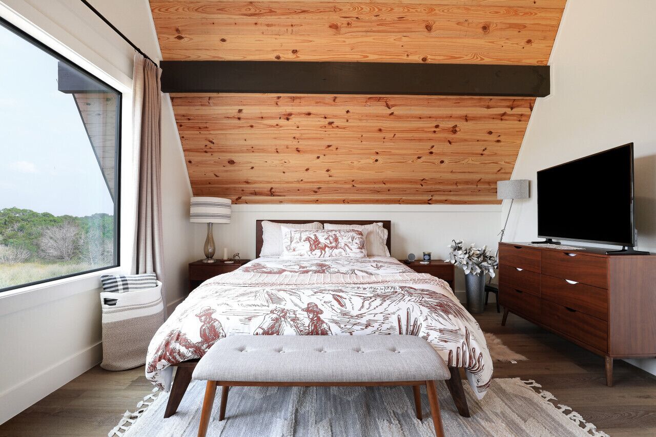 A bedroom with a large bed, wooden ceiling, a TV, and a window with a view.