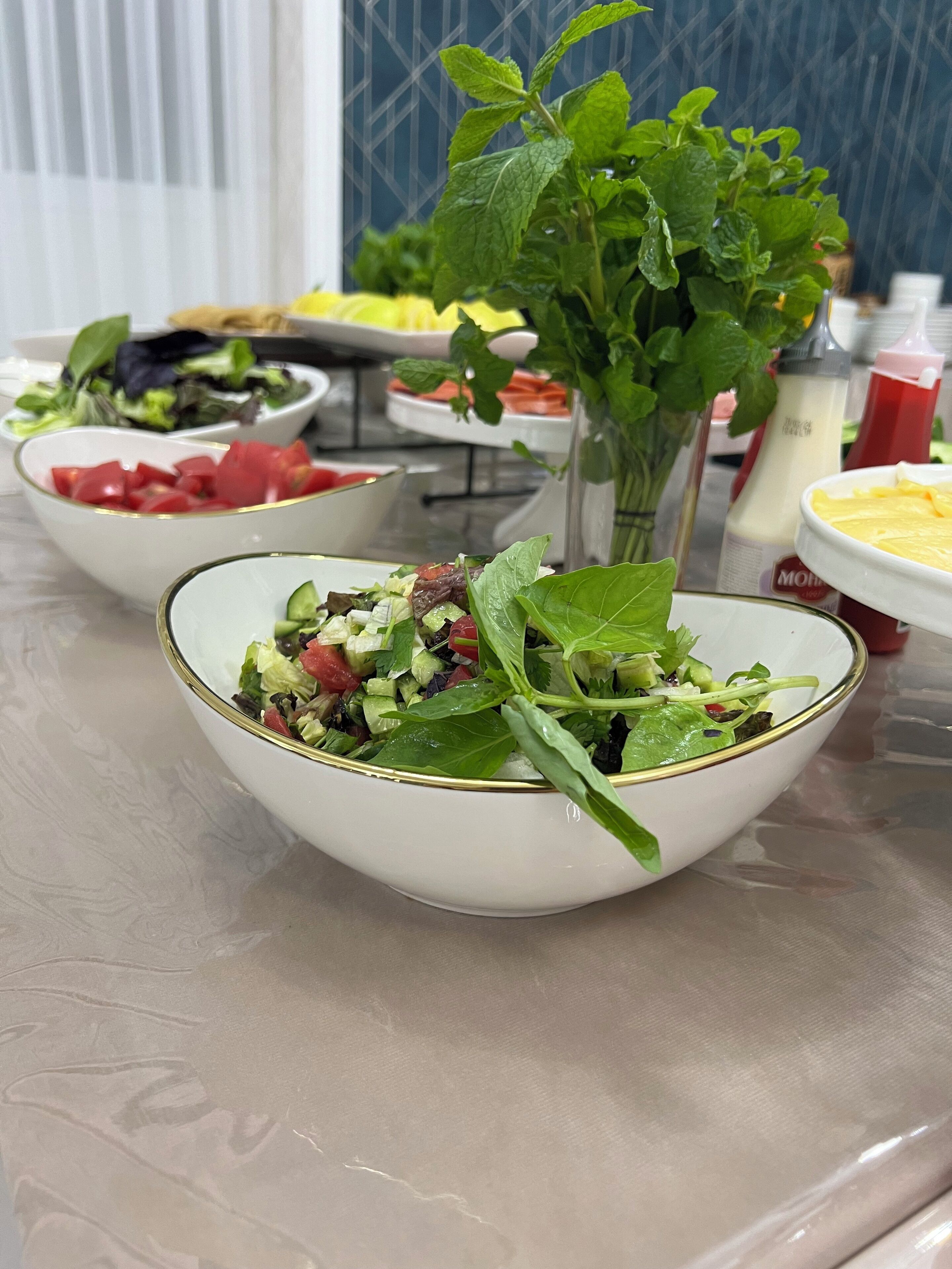 Uma mesa com saladas em tigelas, um vaso com folhas verdes e temperos ao fundo.