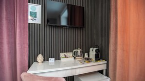 Una habitación moderna con una televisión de pantalla plana montada en la pared, un escritorio blanco con cafetera, un refrigerador pequeño y una silla rosa.