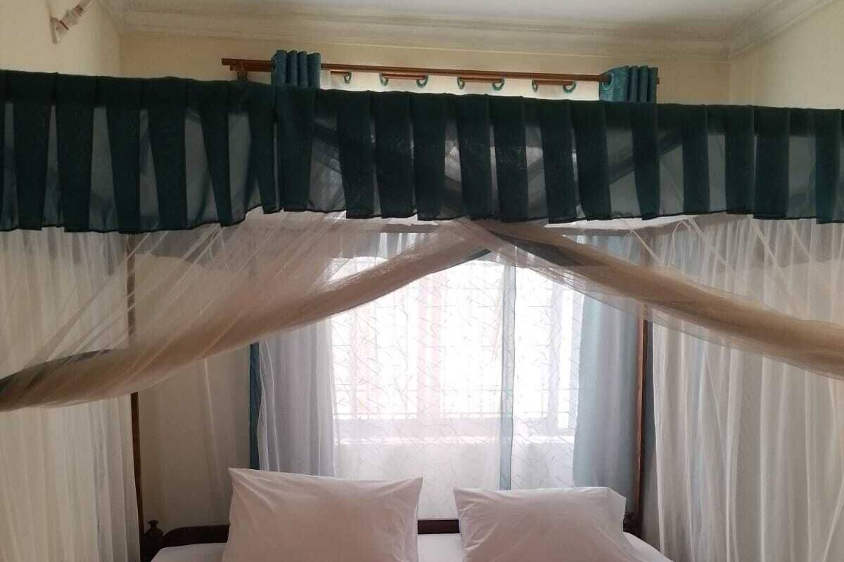 A single bed with white bedding and two pillows, a ceiling fan, and sheer curtains.