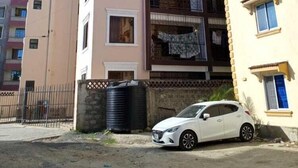 Un edificio residencial de varios pisos con balcones, un coche estacionado frente a él y un depósito de agua cercano.