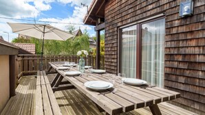 A wooden deck with a picnic table set for four, surrounded by a wooden fence and a house with a large window. - Gorgeous home in Gérardmer with sauna (Gérardmer)