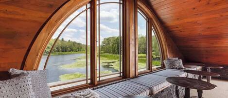 A cozy, wooden interior with a curved ceiling, large windows offering a view of a lake and greenery, a seating area with cushions, and a round table.