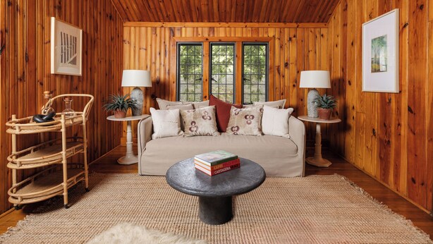 A cozy room with a wooden interior, a sofa, two side tables with lamps, a wicker chair, and a coffee table with books.