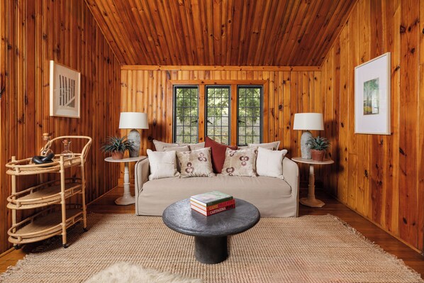 A cozy room with a wooden interior, a sofa, two side tables with lamps, a wicker chair, and a coffee table with books.