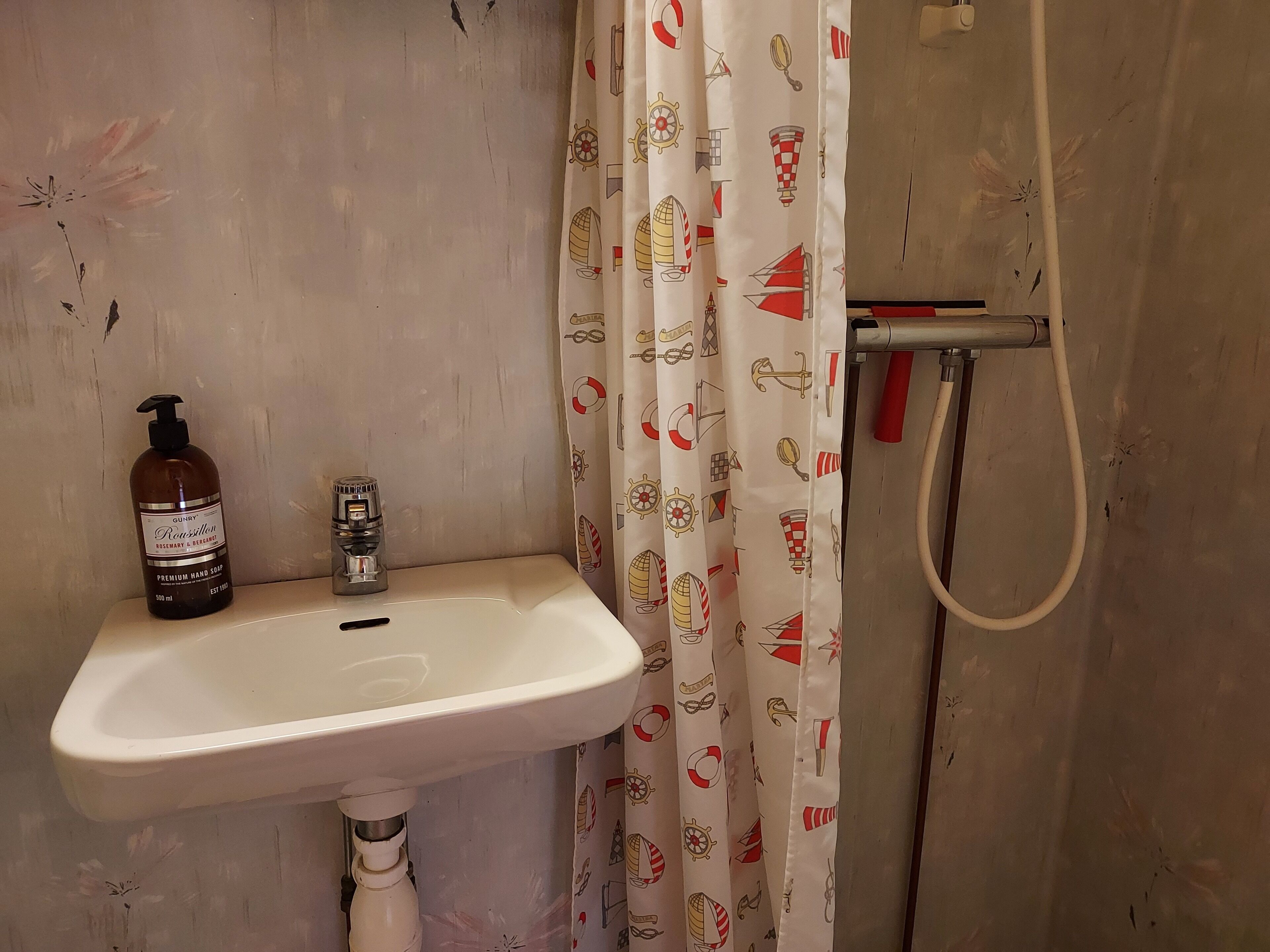 Casa de banho com lavatório branco, um frasco de sabonete e uma cortina de chuveiro com motivos náuticos.