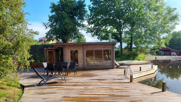 A wooden deck with lounge chairs and a glass-enclosed cabin by a lake. - älgbergets b&b (Ucklum)