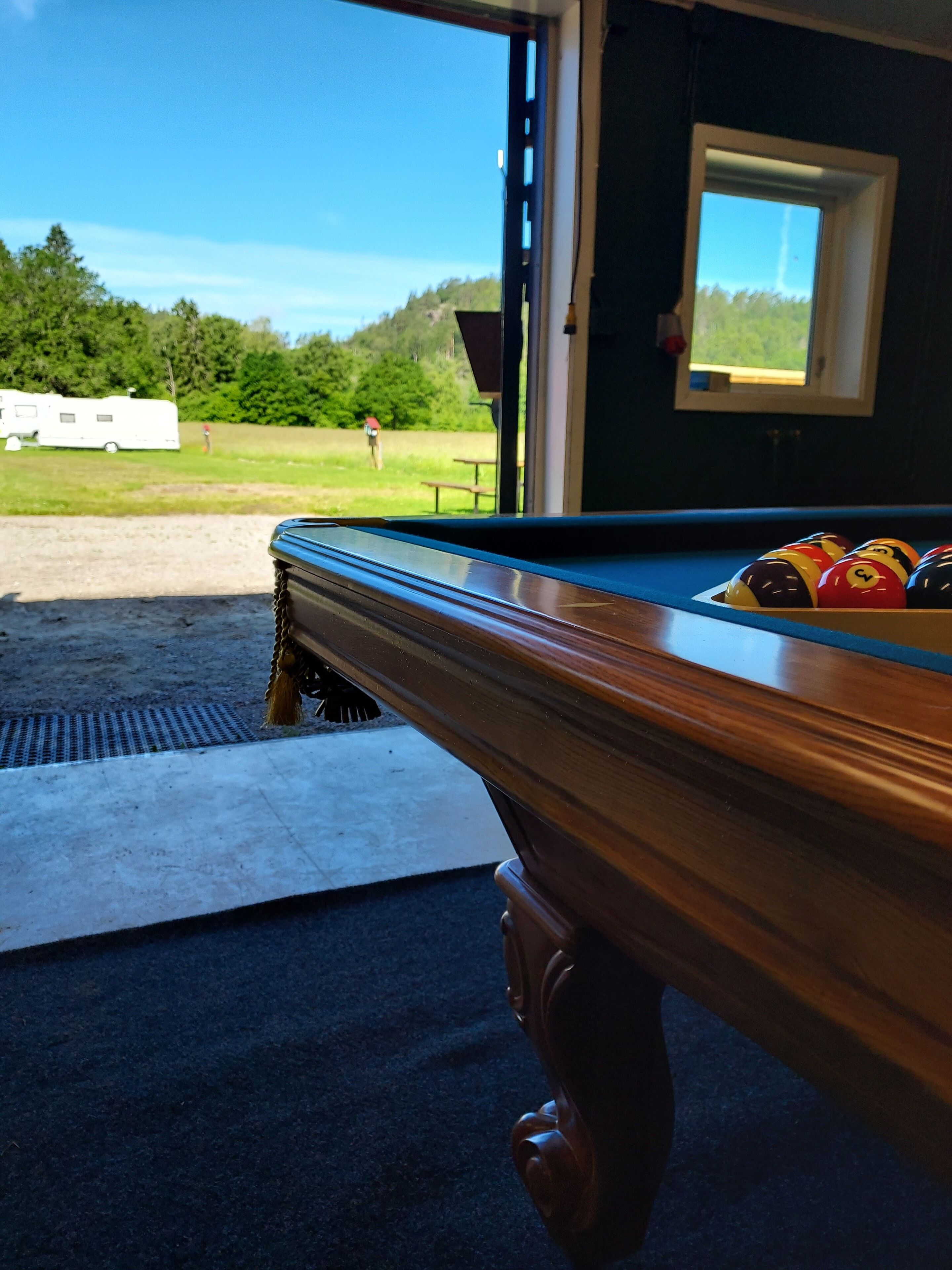 Et carambolebord med kugler, udsigt til en græsplæne og en autocamper i baggrunden.