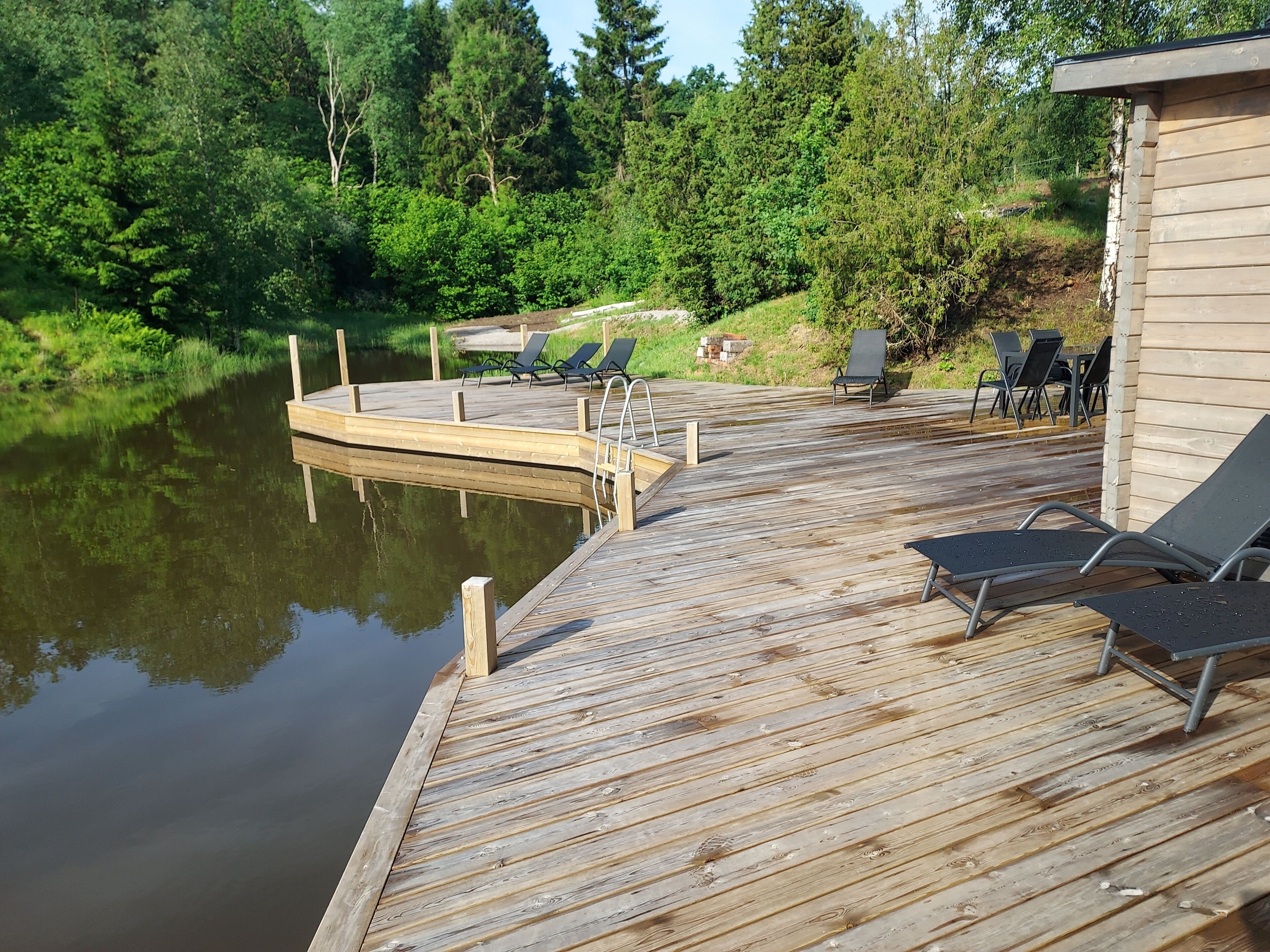 Une terrasse en bois avec des chaises longues et une jetée qui s’avance sur une étendue d’eau calme, le tout entouré de verdure.