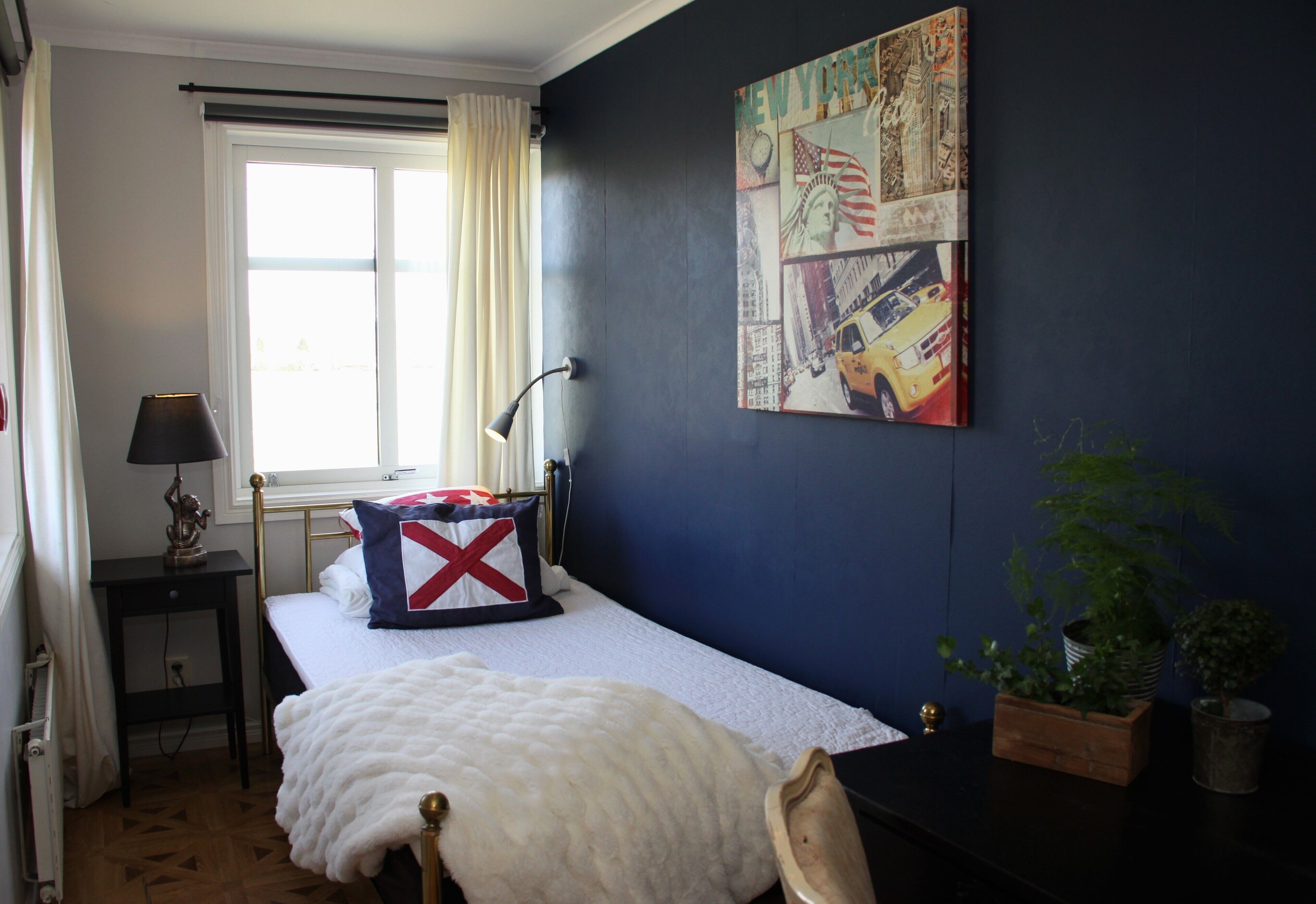 A bedroom with a bed, a nightstand, a lamp, a window with curtains, a wall with a cityscape painting, and a potted plant.