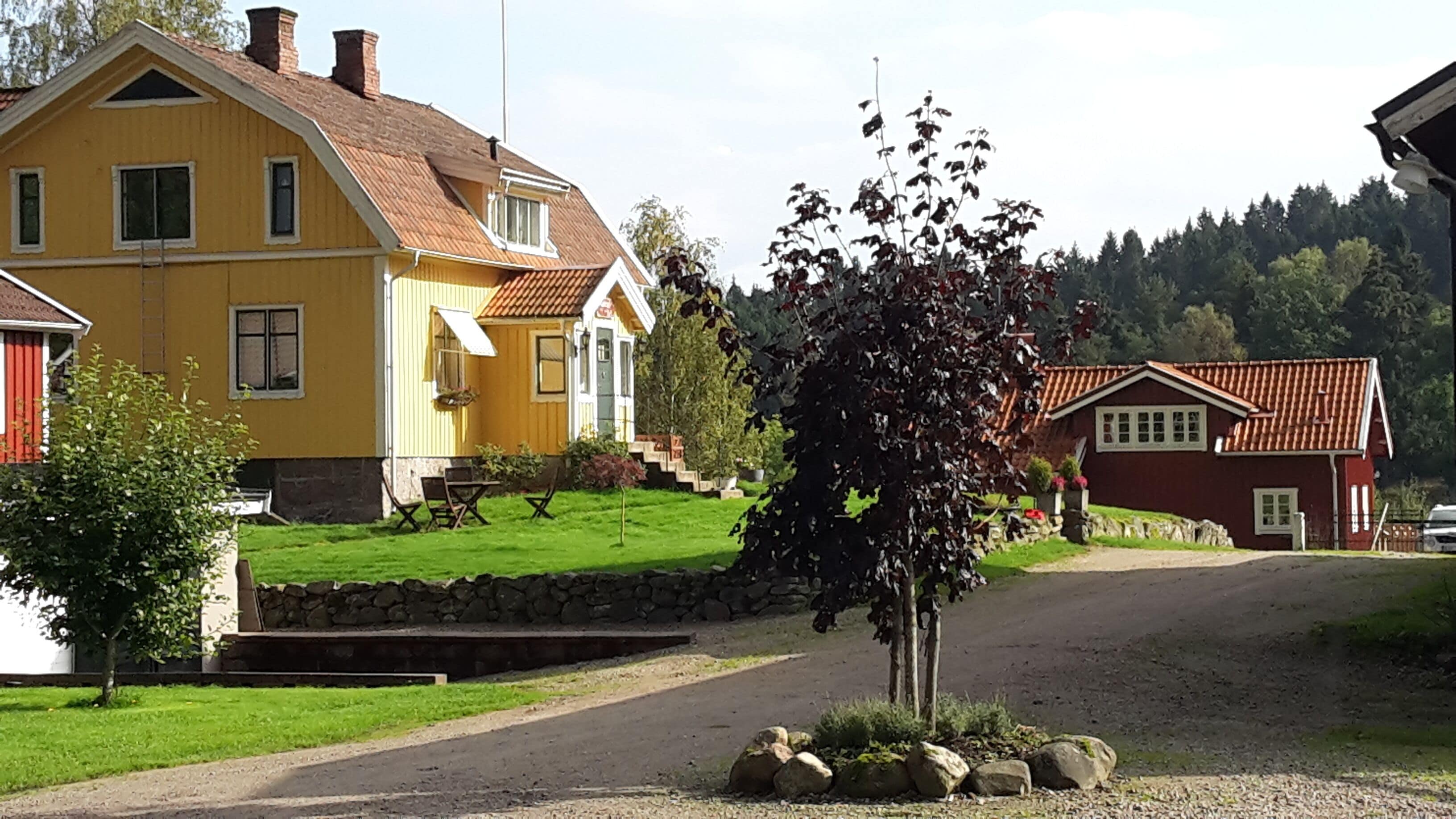 Una casa amarilla con techo rojo, pared de piedra y un pequeño jardín con un árbol.