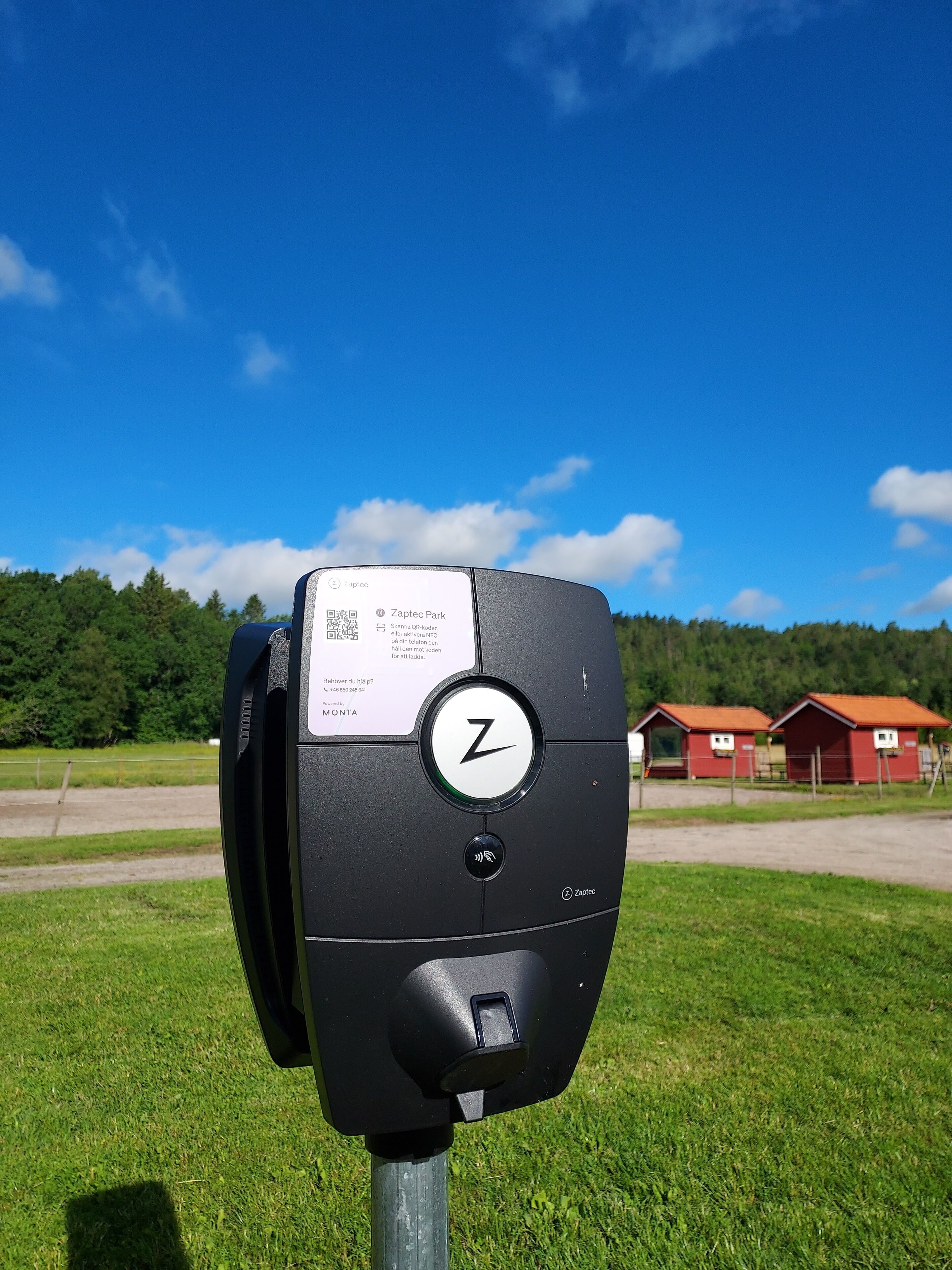 En parkeringsautomat med en QR-kod och en logotyp, placerad mot en lantlig bakgrund med gröna fält och röda byggnader.