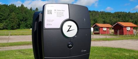 A parking meter with a QR code and a logo, set against a rural backdrop with green fields and red buildings.