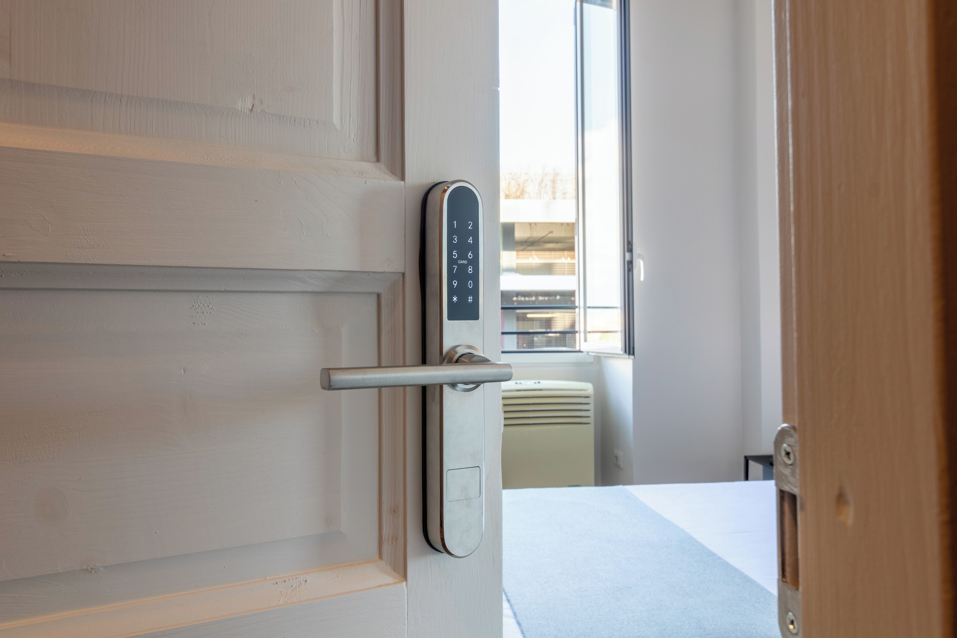 A modern room with a door featuring a numeric keypad lock, a bed with a white and blue bedspread, and a window with a view of buildings outside.