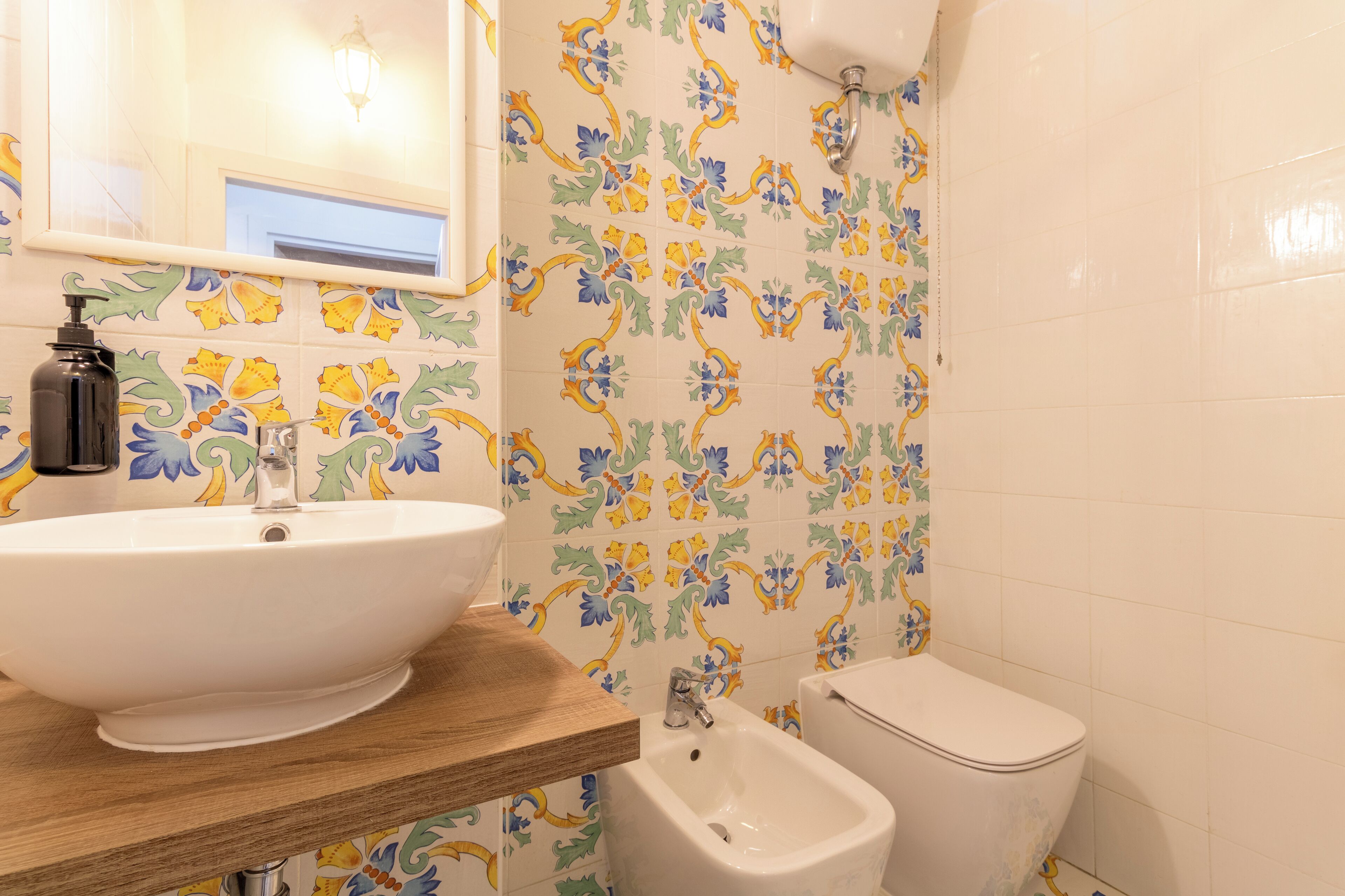 A bathroom with a white sink, wooden countertop, and a bidet. The walls have colorful floral tile patterns.