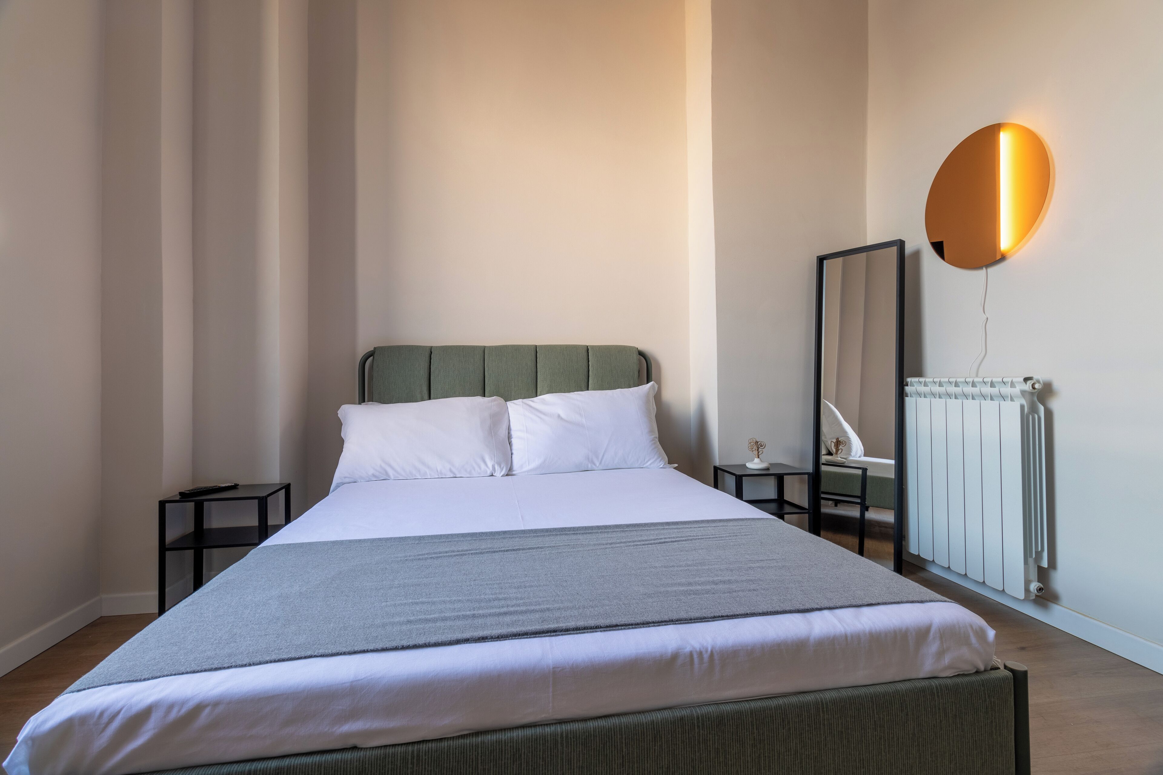 A neatly made bed with white linens and a grey throw, a bedside table with a lamp, and a wall-mounted mirror.