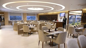 Un restaurante moderno con mesas redondas y asientos acolchados, un área de recepción con televisor y grandes ventanales con vistas a la ciudad.