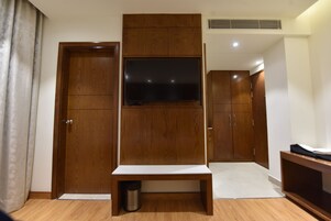 A hotel room with a wooden wardrobe, a television mounted on a wooden panel, a small bench, and a trash bin.