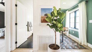 Ein Flur mit Blumentopf, Lüftungsgitter, gerahmtem Bild, Stuhl und einem Fenster mit Vorhängen.