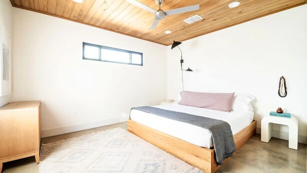 Ein Schlafzimmer mit Holzbalkendecke, einem Bett mit grau-weißer Bettwäsche, einem kleinen Nachttisch mit Lampe und einem Beistelltisch mit Dekoration.