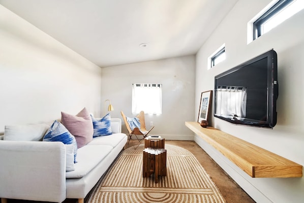 A modern living room with a white sofa, a wooden coffee table, a TV mounted on the wall, and a window with curtains.
