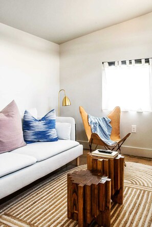 Una sala de estar con un sofá blanco, una mesa de centro de madera, un cojín azul y blanco, un sillón marrón y una lámpara de pie dorada.