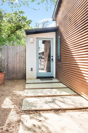 Una casa pequeña y moderna con cerca de madera, un camino de concreto que lleva a una puerta, y una planta en maceta.