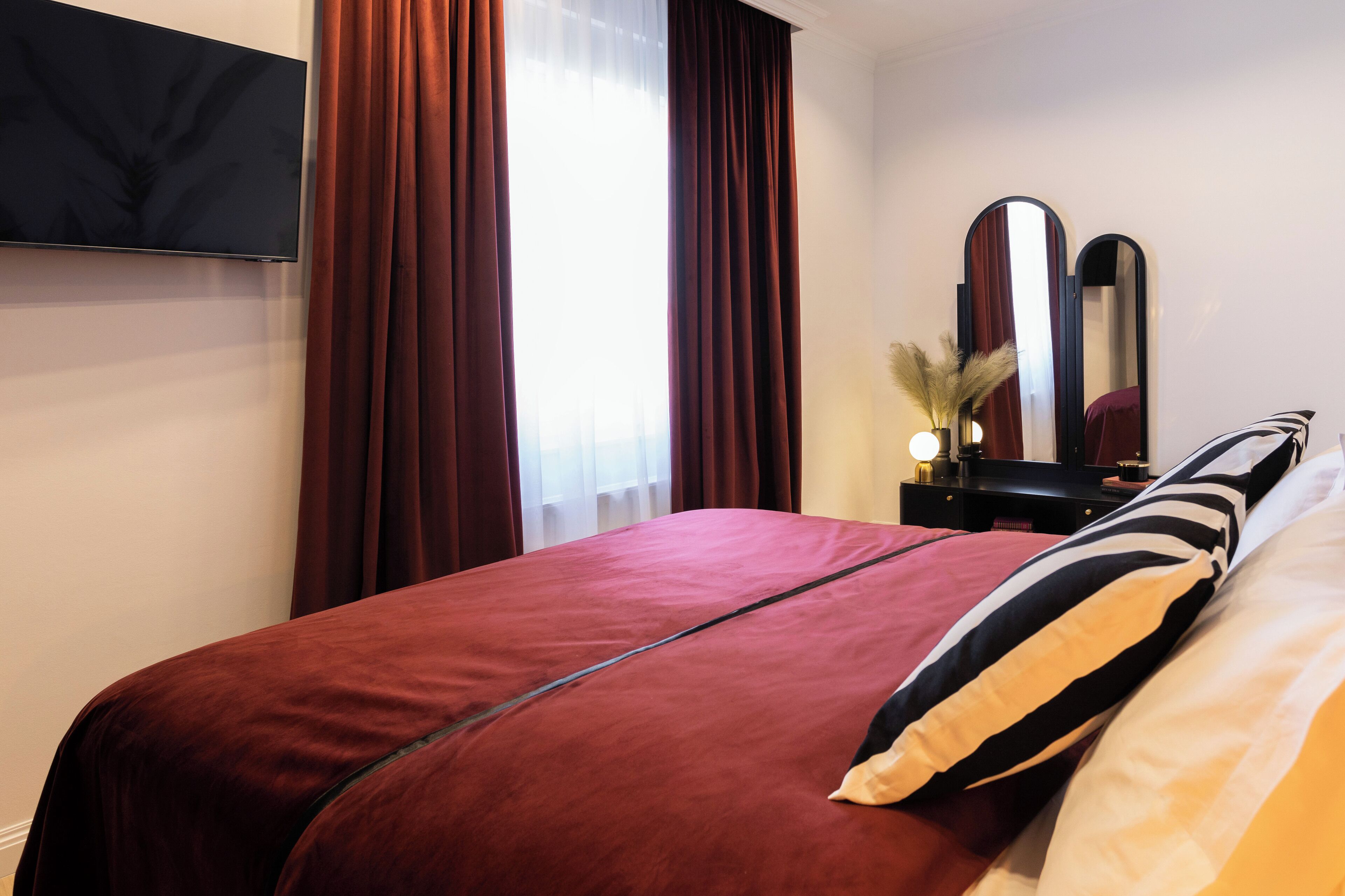 Una habitación de hotel con una cama grande, un televisor de pantalla plana en la pared, un aparador con espejo y una ventana con cortinas.