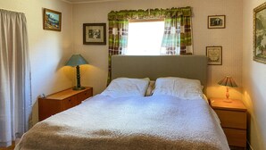 Een netjes opgemaakt bed met een witte dekbedovertrek en twee kussens, met aan weerszijden nachtkastjes met lampen, in een kamer met behang met patroon en afbeeldingen in lijsten aan de wanden.