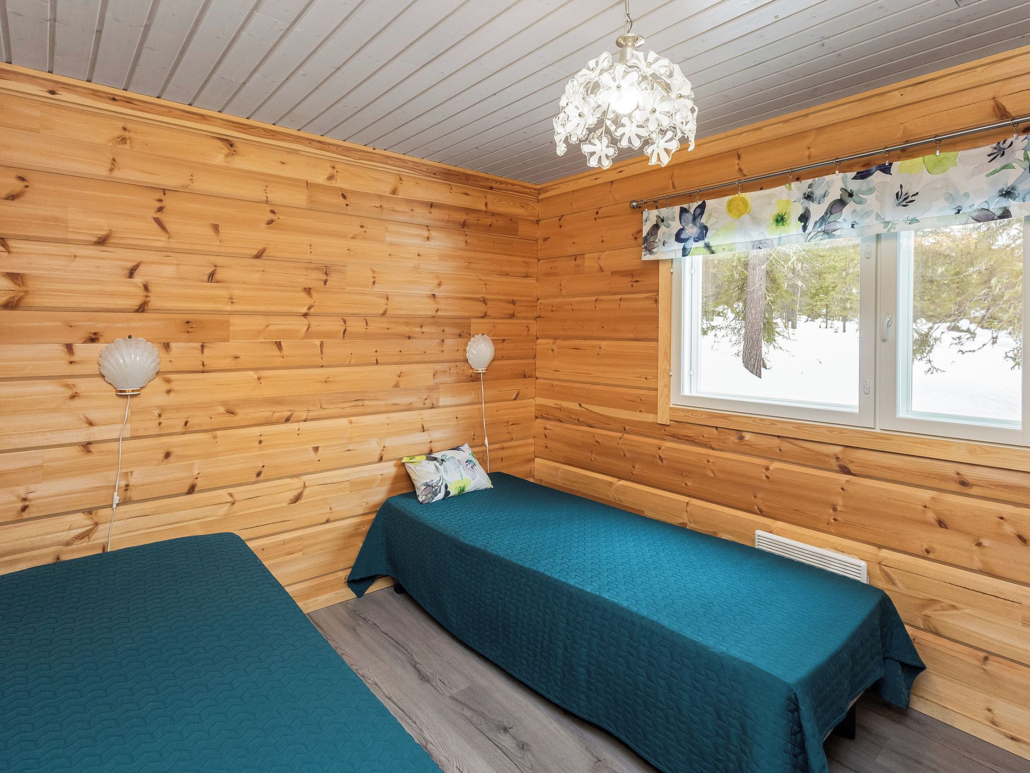 A cabin with two beds, wooden walls, a window with a floral curtain, and a chandelier.