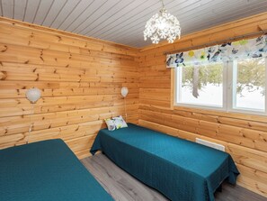 Une cabane avec deux lits, des murs en bois, une fenêtre ornée d’un rideau à motifs floraux et un lustre.