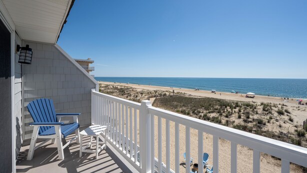 A balcony with a view of the beach, featuring a white railing, two blue Adirondack chairs, and a small table. - Beautiful, modern, 4 bedroom oceanfront townhome! (Ocean City)
