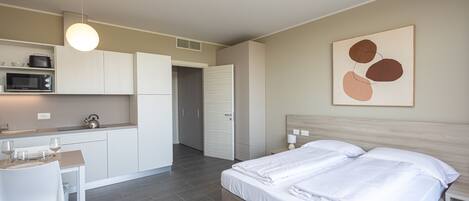 A modern hotel room with a bed, bedside tables, a desk with a chair, a microwave, and a kitchenette.